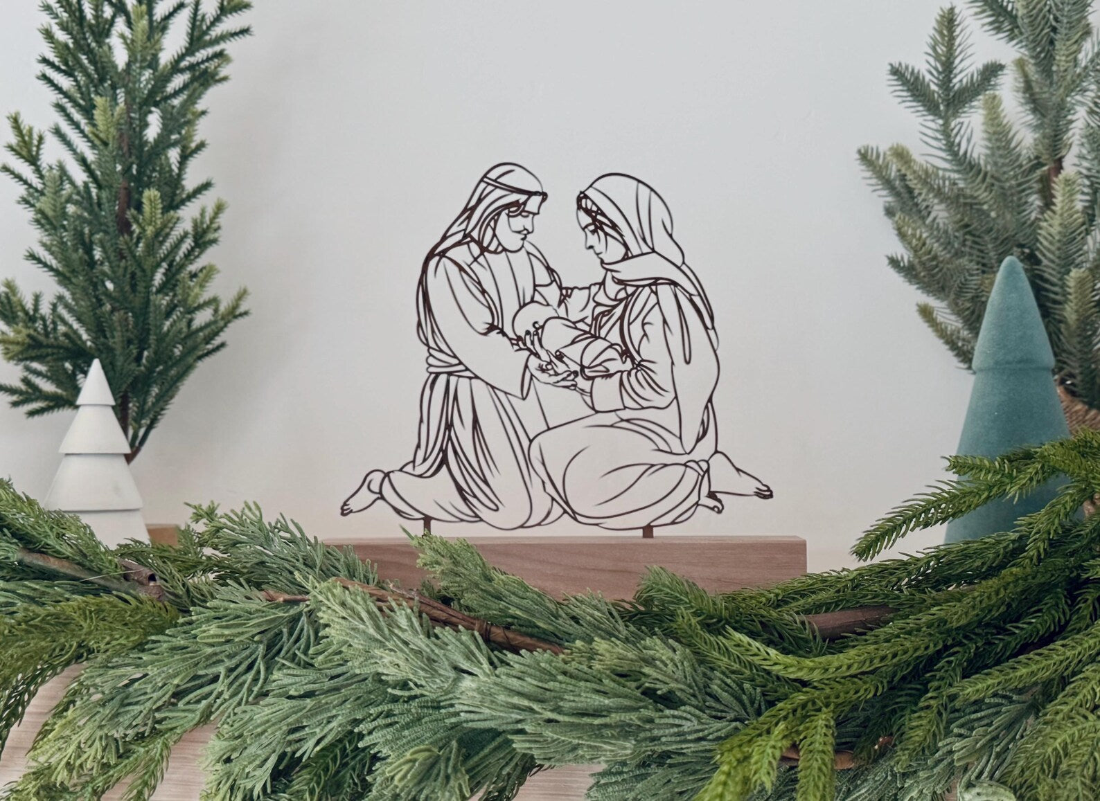 Decorative nativity scene with two figures and a child, surrounded by Christmas greenery.