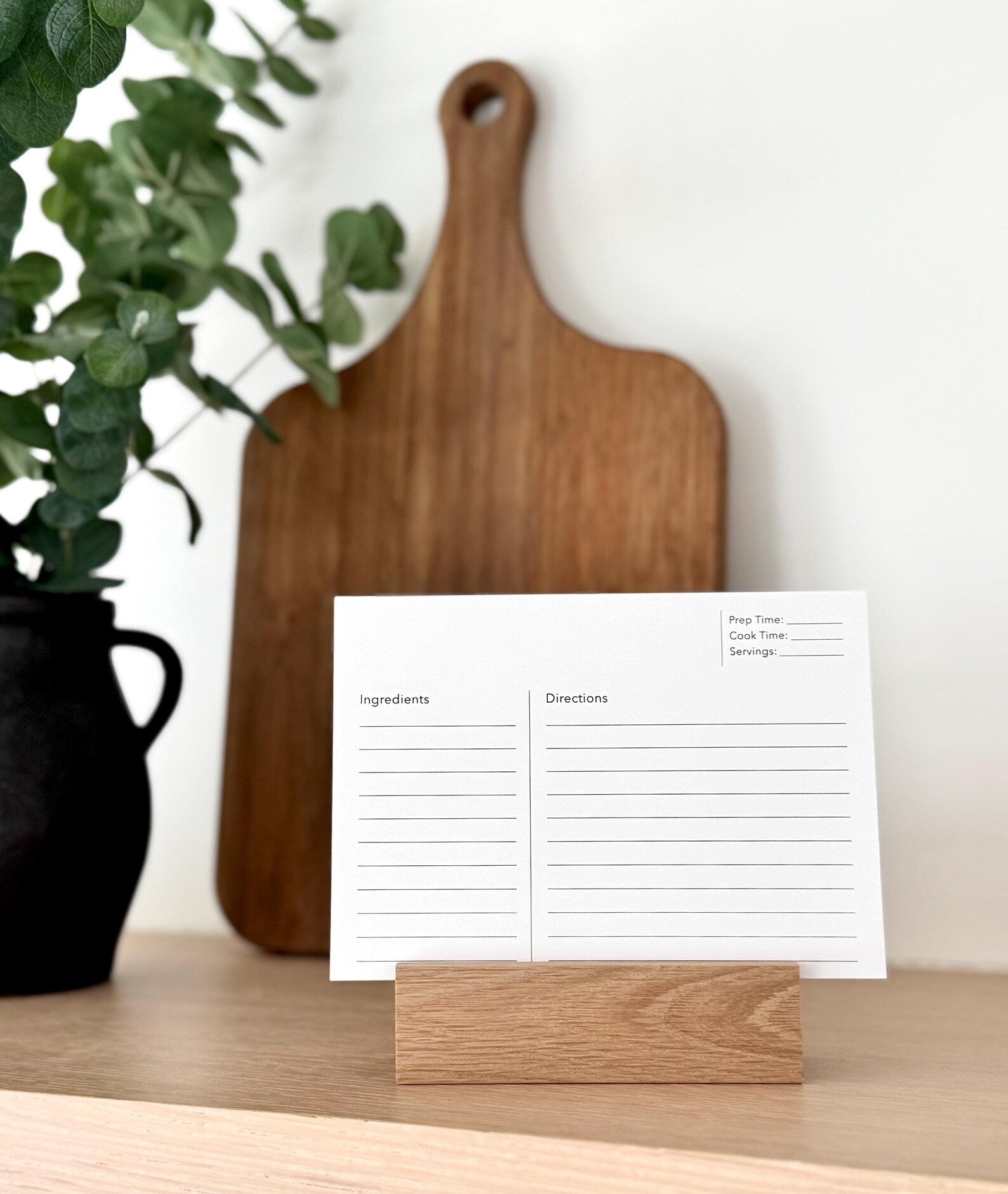 White recipe card with wooden stand on a wooden surface with a plant in the background