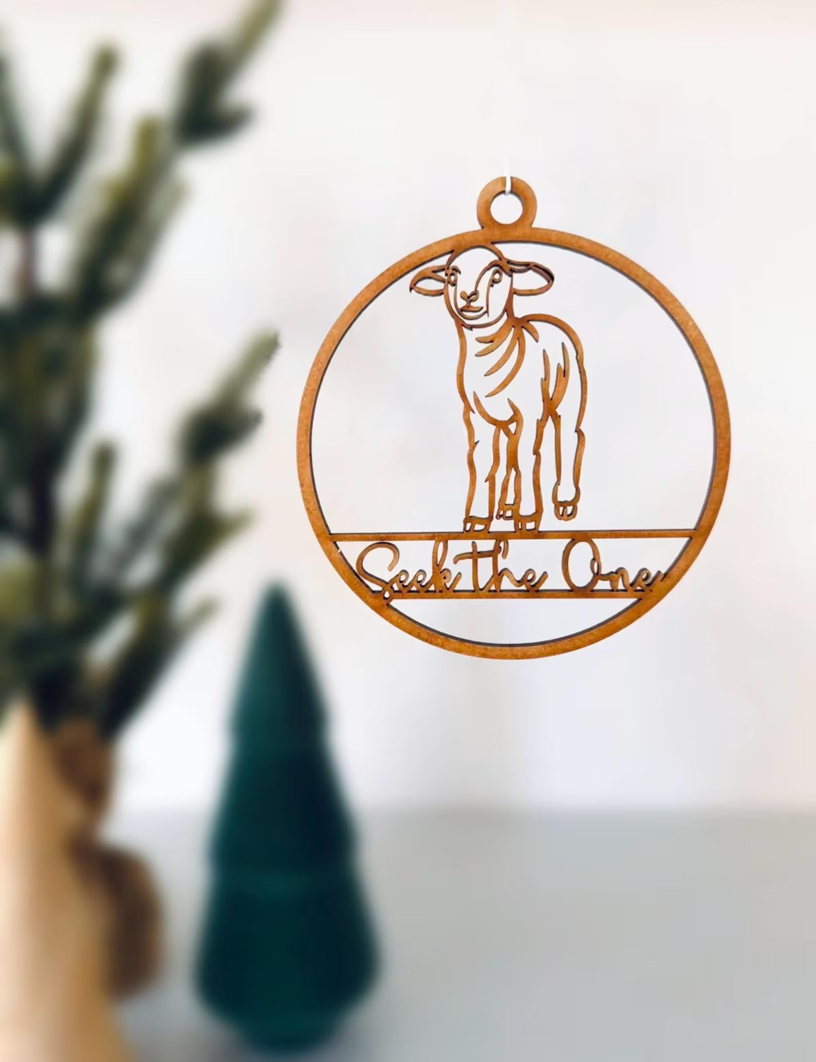 Decorative metal ornament with a sheep design and 'Seek the One' text, placed on a surface with a blurred background.
